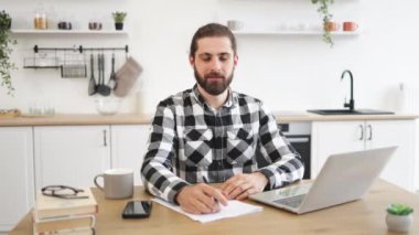 Mutfak arka planında oturmuş kameraya bakarken dizüstü bilgisayar kullanan kareli tişörtlü, kendine güvenen beyaz bir adam. Evde tam zamanlı iş yaparken bilgi arayan verimli bir serbest çalışan..