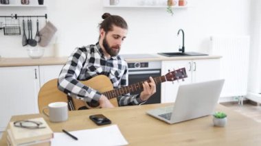 Geniş apartmandaki taşınabilir bilgisayar kamerasına gülümserken elinde telli enstrüman tutan neşeli beyaz insan. Modern teknolojileri kullanarak online gitar dersi veren mutlu bir erkek..