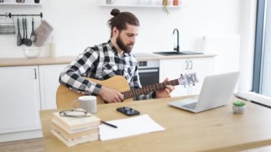 Evde eğlenirken altı telli gitarla poz veren yakışıklı sakallı yetişkin portresi. Gülümseyen beyaz adam bilgisayardan online dersler alarak enstrüman çalmayı öğreniyor..