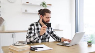 İş yerinde portatif bilgisayar kullanırken kredi kartı kullanan günlük kıyafetler giyen neşeli beyaz bir adam. Kareli tişörtlü, gülen sakallı adam evden gelen online cüzdanı kullanarak sanal alışverişin tadını çıkarıyor.