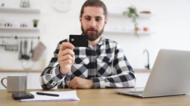 Kafkasyalı mutlu bir beyefendi uzaktan çalışırken online bankacılık işlemleri yapıyor. Kareli tişörtlü çekici sakallı adam elinde kredi kartıyla mutfaktaki taşınabilir bilgisayarda daktilo yazarken..