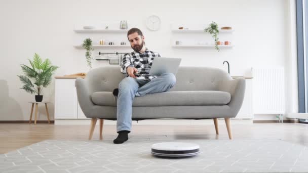 Robotic vacuum removing dust from carpet while Caucasian homeowner utilizing laptop sitting on sofa. Relaxed freelancer leading comfortable lifestyle by using tech gadgets for home kitchen.