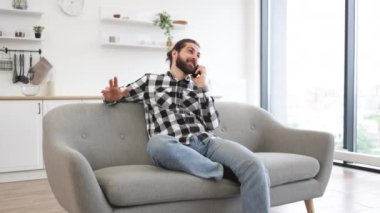 Geniş yemek odasındaki kanepede otururken akıllı telefondan konuşan sıradan giyinmiş neşeli genç adam. Uzaktan kumandalı bir şirketin müşterisiyle verimli bir iş görüşmesi yapan Kafkasyalı serbest çalışan..