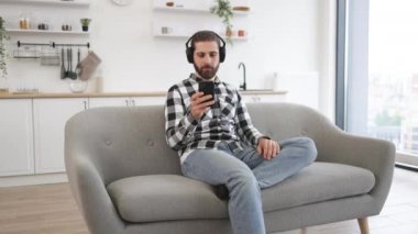 Evde dijital cihazlar üzerinden müzik dinlemeden önce sakin ev sahibi çalma listesini kontrol ediyor. Kafkasyalı rahat bir insan, gündelik giysiler ve kulaklıklar takıyor. Elinde akıllı telefon, mutfakta koltukta oturuyor..