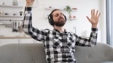 Düğmeli gömlekli neşeli adam mutfakta akıllı telefona bakarken elini kaldırıyor. Mola sırasında kulaklıklarda yüksek sesli melodiler duyarak dans eden neşeli beyaz bir yetişkin..