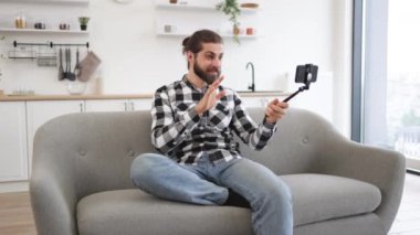 Evdeki koltukta oturan modern bir aletle çevrimiçi konferans düzenleyen mutlu Kafkasyalı. Elinde telefon ve selfie çubuğu olan çekici bir genç adam mutfakta web kamerasıyla konuşuyor..