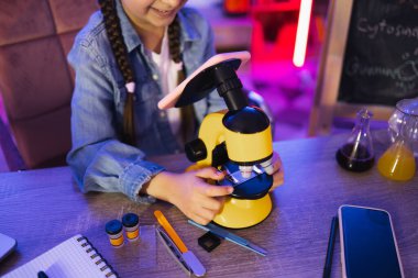 Akşamları evde elektron mikroskobu ve akıllı telefon kullanarak kot gömlekli, fen bilgisi öğrenen odaklanmış küçük kızın ellerine yakından bakın. Çocukluk ve okul kavramını kabul et.