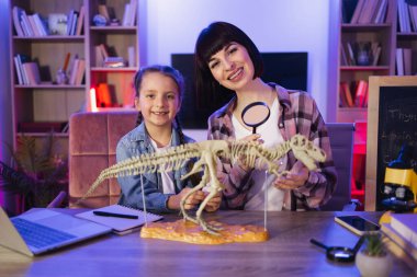 Anne ve kızı evde kemiklerden Tyrannosaurus modeli yapıyorlar. Sıradan giyinmiş genç bir kadın masada küçük bir kızla oturuyor ve büyüteçle dinozorun iskeletini inceliyor..