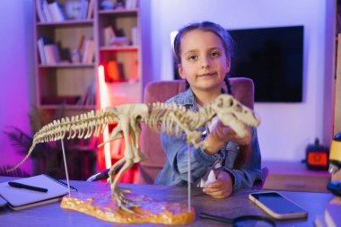 Kot gömlekli küçük kız akşamları evde doğa bilimi öğreniyor kemiklerden tiranozor modeli yapıyor. Çocukluğu ve okulu, ödevi kavramsallaştır.