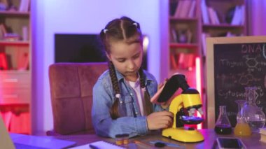 Kot gömlekli küçük kız evde elektron mikroskobu ve akıllı telefon kullanarak fen bilgisi öğreniyor. Çocukluk ve okul kavramını kabul et.