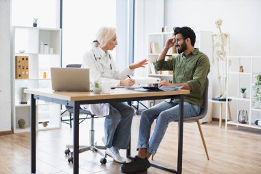 Yaşlı kadın doktor, hastane ofisindeki genç hastaya uzman kontrolü ve reçete yazıyor. Baş ağrısı olan bir adam tıbbi geçmişiyle ilgili tavsiyeleri dikkatle dinliyor..