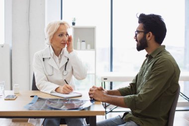 Olgun bir doktor tavsiye veriyor ve reçete yazmak için not defteri kullanıyor. Yaşlı kadın doktorlara hastalık belirtileri gösteren sakallı bir beyefendi..