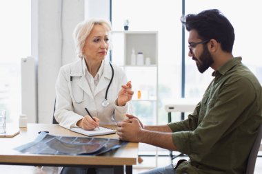 Sakallı beyefendi, yaşlı kadın doktora hastalık belirtilerini ve el kol hareketlerini anlatıyor. Olgun bir doktor tavsiye veriyor ve reçete yazmak için not defteri kullanıyor..