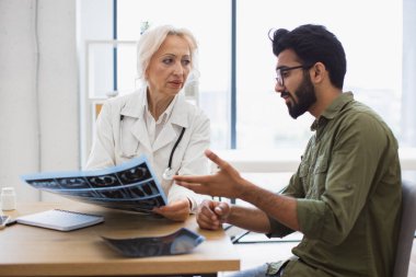 Aile doktoru tedavi planını geliştirirken teşhis sonuçlarını inceliyor. Yanında oturan esmer adam hastanede masasında otururken, elinde MRI taraması tutan, laboratuvar önlüklü, beyaz saçlı dikkatli bir kadın..