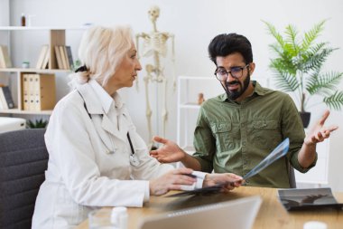 Beyaz üniformalı yetenekli radyolog röntgen görüntülerindeki gözle görülür değişiklikleri açıklıyor. Günlük giysiler giymiş endişeli bir adam doktorların talimatlarını dikkatle dinliyor..