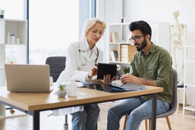 İlaçları doğru almak için gerekli elektronik talimatları okuyan yetişkin bir uzman. Kıdemli kadın doktor ve erkek hasta modern tıp merkezinin masasında oturup tablette birlikte izliyorlar..