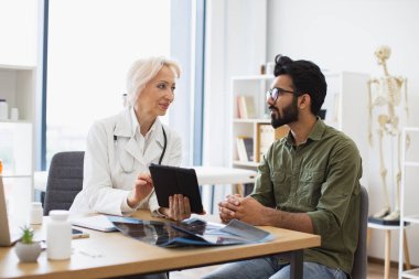 Kadın doktora hastalık belirtilerini tarif eden genç bir beyefendi. Gri saçlı doktor tavsiye veriyor ve elektronik reçete yazmak için tablet kullanıyor..