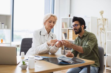 Sakallı beyefendi, yaşlı kadın doktora hastalık belirtilerini ve vücut ısısını tarif ediyor. Olgun yaşlı doktor tedaviyle ilgili tavsiyelerde bulunuyor.