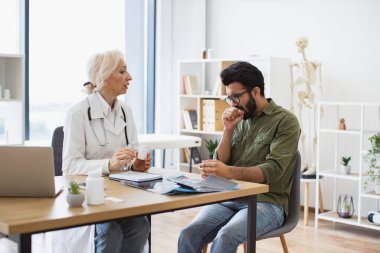 Yaşlı kadın doktor, hastane ofisindeki genç hastalara uzman muayenesi ve ilaç veriyor. Bilgisayarda tutulan tıbbi geçmişle ilgili tavsiyeleri dikkatle dinleyen odaklanmış bir adam..