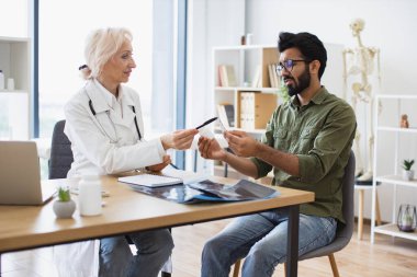 Kıdemli kadın pratisyen doktor yeni klinikte elinde şişeyle genç bir hastayla iletişim kuruyor. Doktor sağlık sorunlarını tartışıyor ve laptoptaki tıbbi verileri inceliyor..