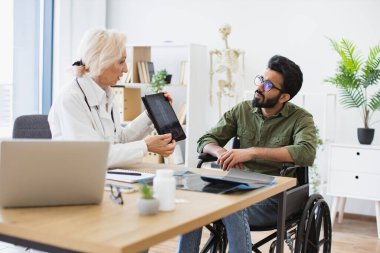 Etkili bir travmatolog hastanenin iç kısımlarındaki teşhisler konusunda hastaya güven veriyor. Beyaz önlüklü yaşlı kadın tablet üzerinde omurga taraması yapıyor. Tekerlekli sandalye kullanan dikkatli bir adama..