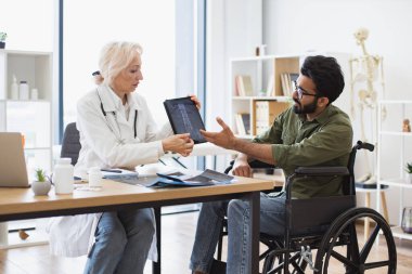 Etkili bir travmatolog hastanenin iç kısımlarındaki teşhisler konusunda hastaya güven veriyor. Beyaz önlüklü yaşlı kadın tablet üzerinde omurga taraması yapıyor. Tekerlekli sandalye kullanan dikkatli bir adama..