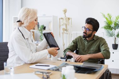 Radyologlar tarafından danışma odasında taşınan röntgen görüntüsüyle tablete bakan çekici sakallı genç adam. Tekerlekli sandalye kullanıcısıyla teşhis testlerinin sonuçlarını tartışan kendinden emin uzman.