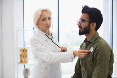 Her gün ciddi sakallı bir adam genel muayene için aile doktorunu ziyaret ediyor. Beyaz önlüklü yaşlı kadın steteskopla akciğer ve kalbi kontrol ediyor..