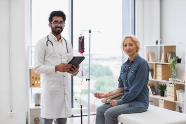 Laboratuvar önlüğü giymiş deneyimli bir adam, kanepede oturan yaşlı kadının kan naklini izlemek için tablet kullanıyor. Klinikte kanser olan kadın hastalara danışmanlık yapan bir sağlık uzmanı..