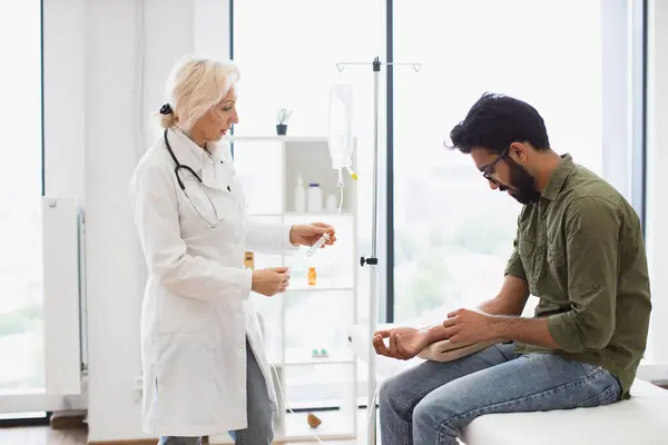 Young bearded man in casual wear taking dropper while mature female making system review. Efficient general practitioner checking solution for chemotherapy of male sitting on exam couch.