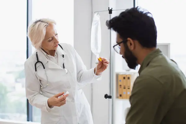 Medical specialist checking drug solution before dripping medicine of patient in exam room. Bearded young man receiving chemotherapy looking at nurse in lab coat preparing a dropper.