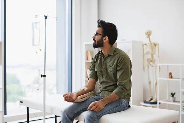 Young bearded man taking dropper with solution for chemotherapy sitting on exam couch. Tired sick male in casual wear drips medicine intravenously while sitting on modern light ward.