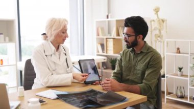 Beyaz üniformalı yetenekli radyolog röntgen görüntülerindeki gözle görülür değişiklikleri açıklıyor. Günlük giysiler giymiş endişeli bir adam doktorların talimatlarını dikkatle dinliyor..