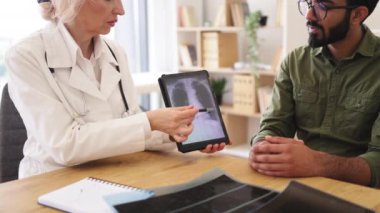 Beyaz üniformalı yetenekli radyolog röntgen görüntülerindeki gözle görülür değişiklikleri açıklıyor. Günlük giysiler giymiş endişeli bir adam doktorların talimatlarını dikkatle dinliyor..