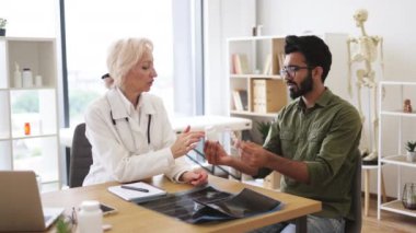 Doktor sağlık sorunlarını tartışıyor ve laptoptaki tıbbi verileri inceliyor. Kıdemli kadın pratisyen doktor yeni klinikte elinde şişeyle genç bir hastayla iletişim kuruyor..