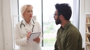 Sakallı beyefendi, yaşlı kadın doktora hastalık belirtilerini ve el kol hareketlerini anlatıyor. Olgun bir doktor tavsiye veriyor ve reçete yazmak için not defteri kullanıyor..