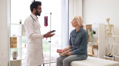 Laboratuvar önlüğü giymiş deneyimli bir adam, kanepede oturan yaşlı kadının kan naklini izlemek için tablet kullanıyor. Klinikte kanser olan kadın hastalara danışmanlık yapan bir sağlık uzmanı..