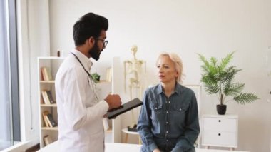 Orta dereceli tıp bürosundaki genç erkek doktora hastalık belirtilerini tarif eden yaşlı bir kadın. Yetişkin sakallı doktor tavsiye veriyor ve elektronik reçete yazmak için tablet kullanıyor..