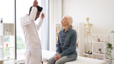 Yaşlı bir kadın muayene koltuğunda otururken beyaz önlüklü, steteskop kullanan odaklanmış yetişkin bir adamın yan görüntüsü. Klinikte düzenli kontrol sağlarken kalp atışlarını inceleyen pratisyen hekim.