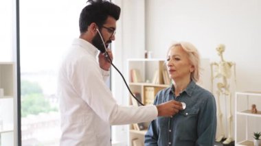 Klinikte düzenli kontrol sağlarken kalp atışlarını inceleyen pratisyen hekim. Yaşlı bir kadın muayene koltuğunda otururken beyaz önlüklü, steteskop kullanan odaklanmış yetişkin bir adamın yan görüntüsü..