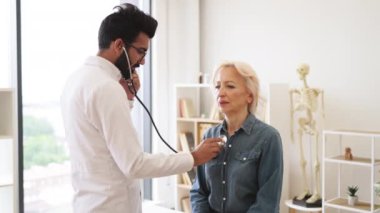 Beyaz laboratuvar önlüklü sakallı adam steteskopla akciğer ve kalbi kontrol ediyor. Ciddi yaşlı bir kadın günlük hayatta, aile doktorunu muayene ederken öksürüyor..