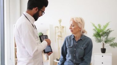 Aile doktoru tedavi planını geliştirirken teşhis sonuçlarını inceliyor. Yanında yaşlı bir kadın otururken elinde MRI taraması olan laboratuvar önlüklü, dikkatli sakallı bir erkek..