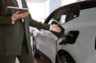 Beyaz tenli genç bir satıcının, panoramik pencereli, modern, aydınlık sergi salonunda elektrikli araba satışı görüntüsü. İlerici müşteriler için çevre dostu araba alma kavramı.