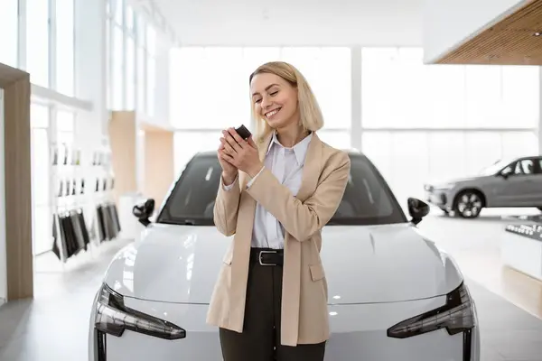 stock image Happy smiling business lady standing near new bought car and hugging keys in car dealership. Successful girl in suit rejoices in a new car. Trading concept.