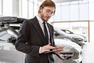 Gözlüklü ve takım elbiseli genç araba satıcısı dijital tablet tutuyor. Kendine güvenen sakallı bir adam modern aletlerle yeni bir araba satın almak için emir verir..