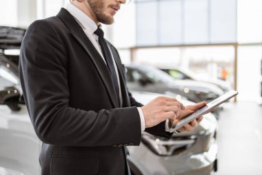 Elinde dijital tablet olan takım elbiseli genç araba satıcısının görüntüsü. Kendine güvenen sakallı adam modern aletlerle yeni bir araba almak için emir veriyor..