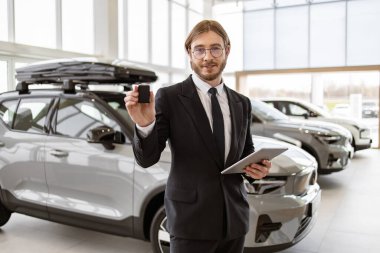 Kendine güvenen satıcı, galerideki yeni arabanın anahtarını müşteriye veriyor. Araba satıcısı neşeyle gülümsüyor, anahtarı tutuyor, satılık yeni araçların önünde duruyor. Teklif, indirim kavramı.