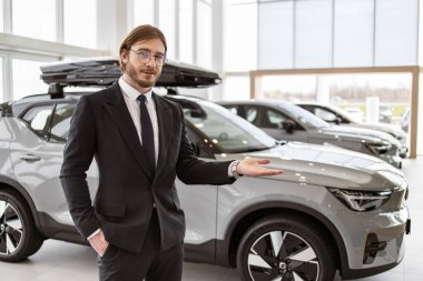 Gülümseyen beyaz satıcı kameraya bakıyor ve sergi salonundaki yeni araba modellerini işaret ediyor. Araba galerisinde gururla poz veren takım elbiseli ve gözlüklü profesyonel sakallı bir araba satıcısı..