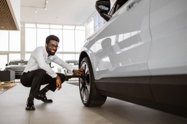 Müşteri, alıcı, beyaz tişörtlü müşteri otomobili seçiyor. Gülümseyen işadamı, galeride araba lastiği diskine dokunurken yeni bir otomobil almak istiyor..