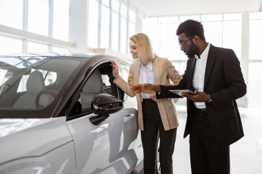 Araba galerisinde müşterilere araç satışında başarılı bir iş kadını. Afrikalı erkek danışman, araba galerisindeki beyaz kadın müşteriye tablet kullanarak yeni araba hakkında bilgi sağlıyor.